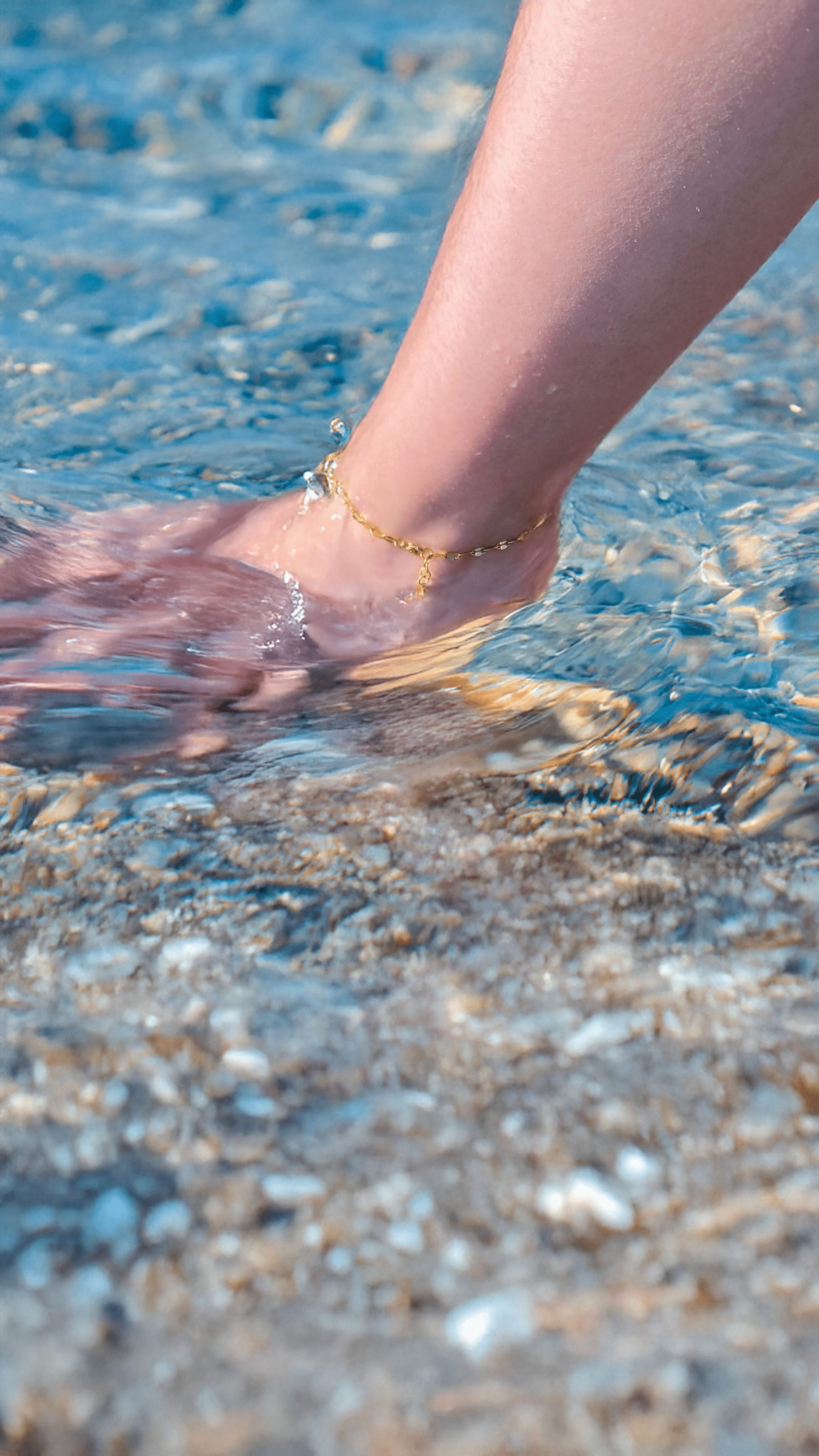 A soft, gold-tone chain with a central CZ stone is shown elegantly resting on skin, styled as both anklet and bracelet. It shimmers as the model moves gently, highlighting the chain’s flexibility and detail. Anklet in water.